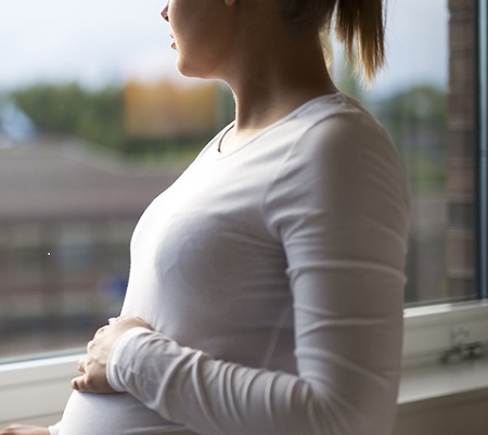 Thoughtful pregnant woman looks out the window home. Holding a hand on the tummy. Mental health and pregnancy.