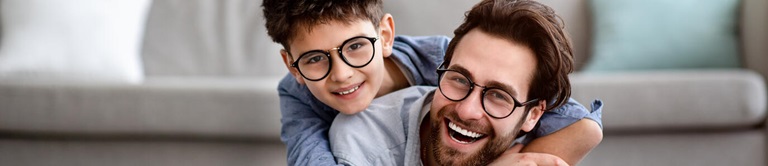 Son smiling and hugging his dad in on the living room ground with the couch in the background, both wearing glasses